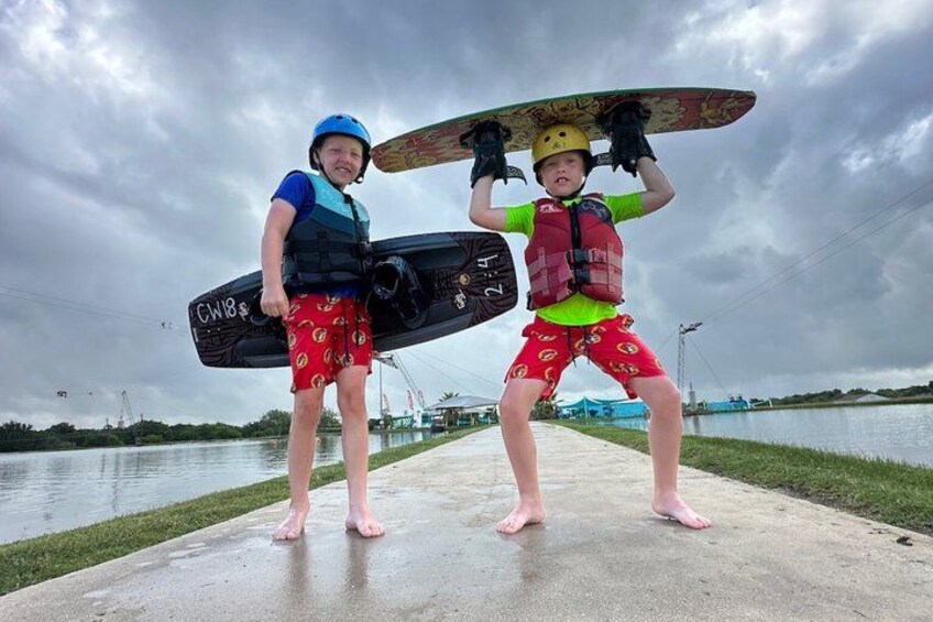 Two people holding wakeboards on a concrete path near a body of water. 1