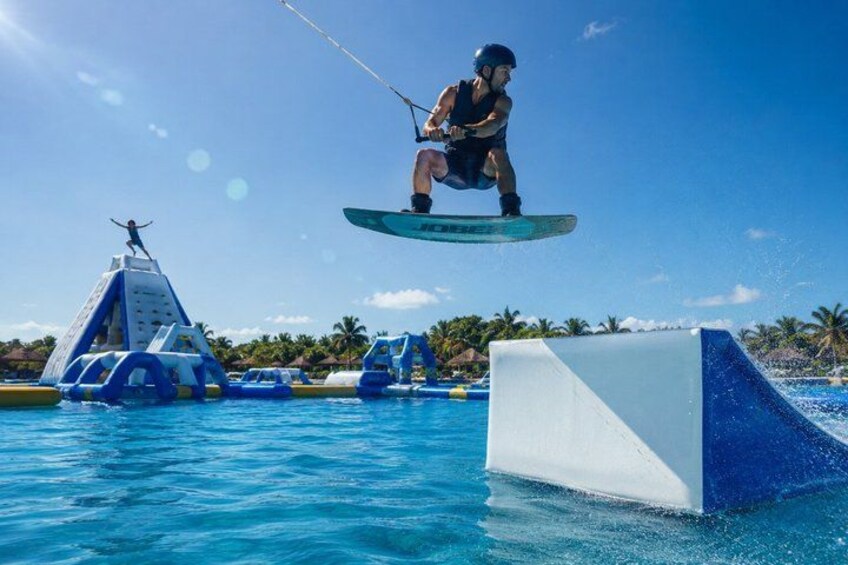 A person wakeboarding over a pool with inflatable structures and palm trees in the background. 2