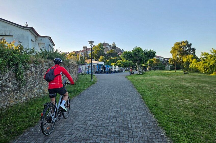 A cyclist on a paved path with grass on the sides, a building on the left, and trees in the background. 3