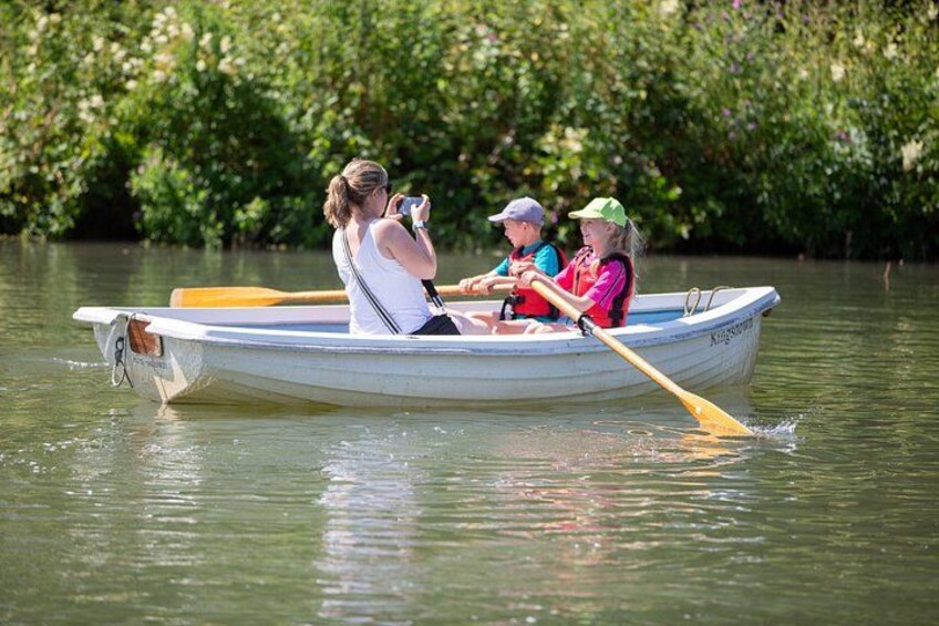 A rowboat with three people rowing on a calm river surrounded by dense greenery. 3