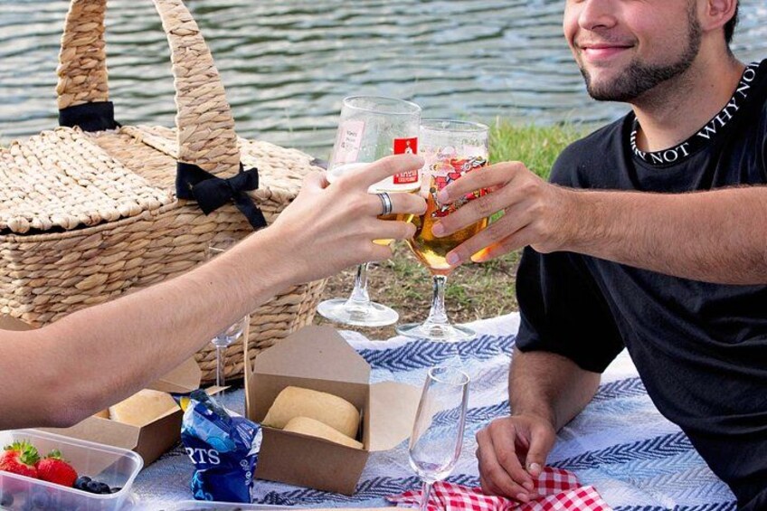 Two people toasting with wine glasses at a lakeside picnic with a basket and snacks. 4