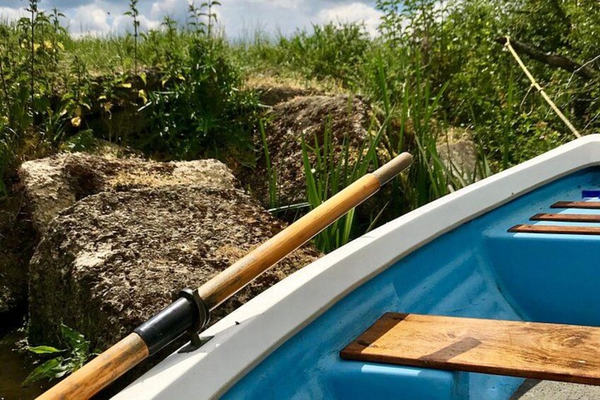 A rowboat with wooden oars and a blue interior, docked near a rocky shore with vegetation. 1