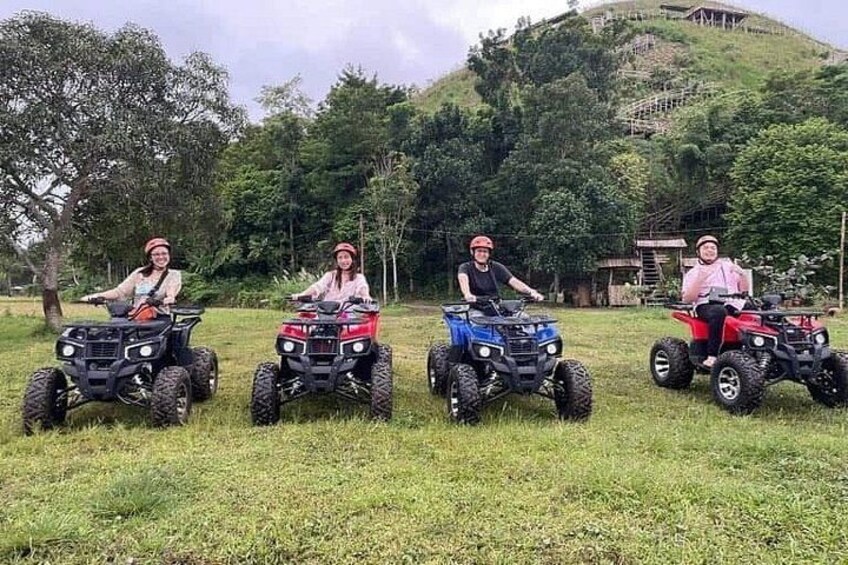 Three people on quad bikes in a grassy field with trees and a hill in the background. 4