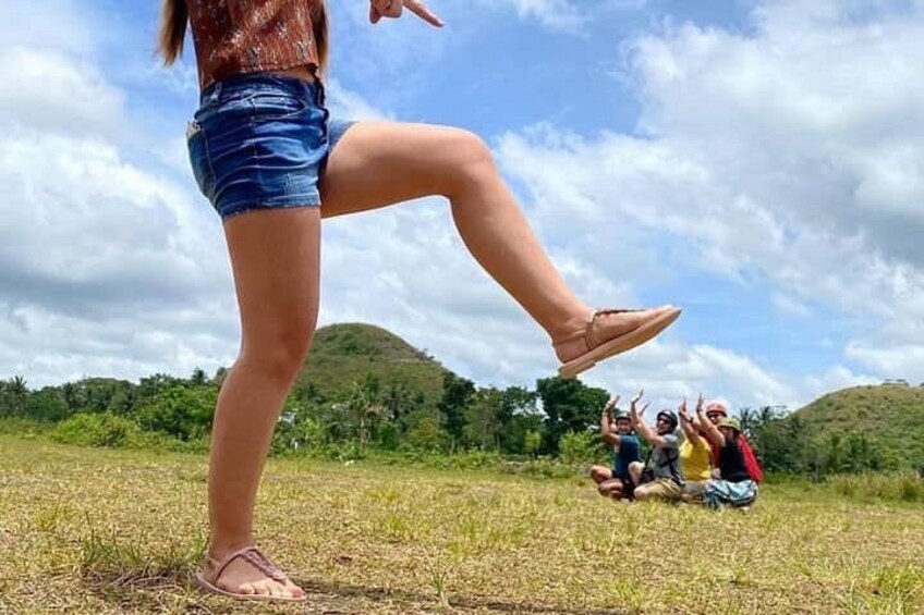 A person standing with one leg raised, a group of people sitting on the ground in the background, and a hilly landscape with trees. 5