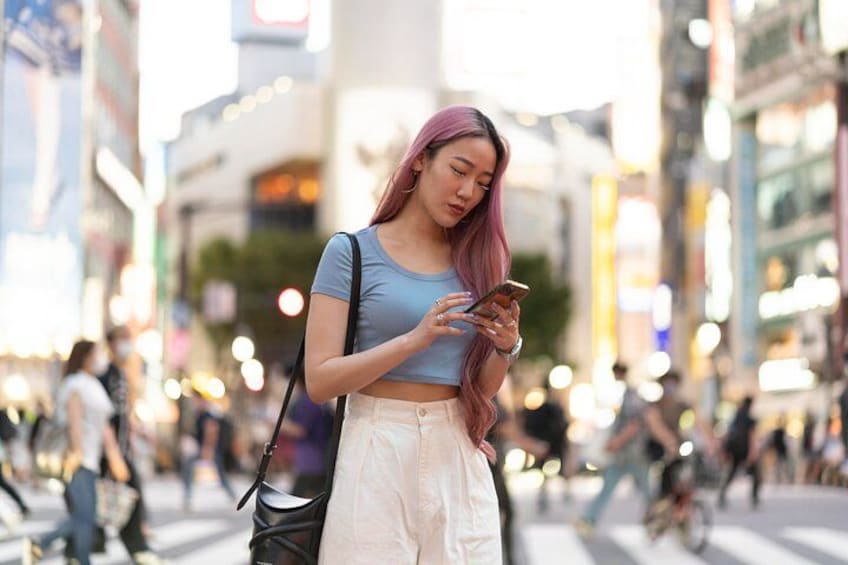 A person with pink hair standing on a busy street, holding a smartphone. 3