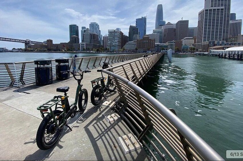 A waterfront walkway with bicycles parked along the railing, a city skyline in the background, and a bridge crossing the water. 3