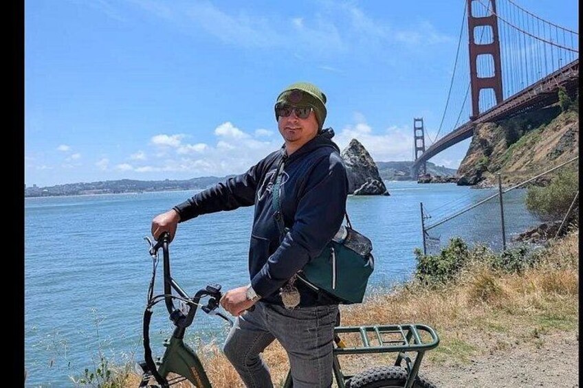 A person with a bicycle and a green bag near a body of water with a bridge in the background. 5