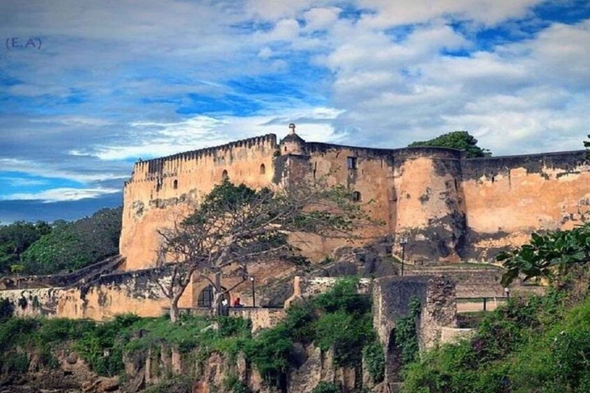 Una gran fortaleza de piedra histórica con una torre destacada, rodeada de abundante vegetación y un cielo despejado. 1