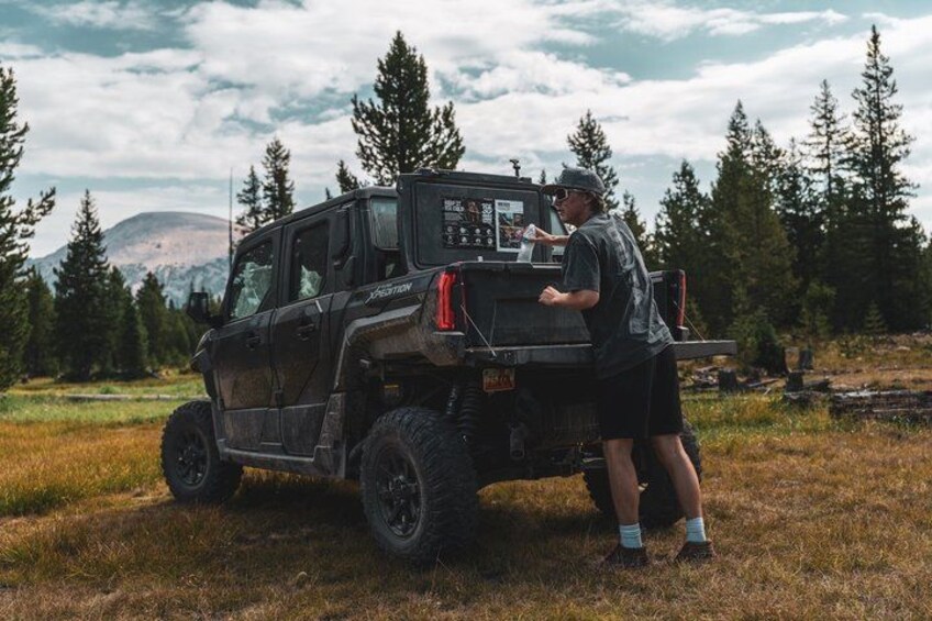 A black off-road vehicle with a mounted winch and a person standing beside it in a grassy field with trees and mountains in the background. 1