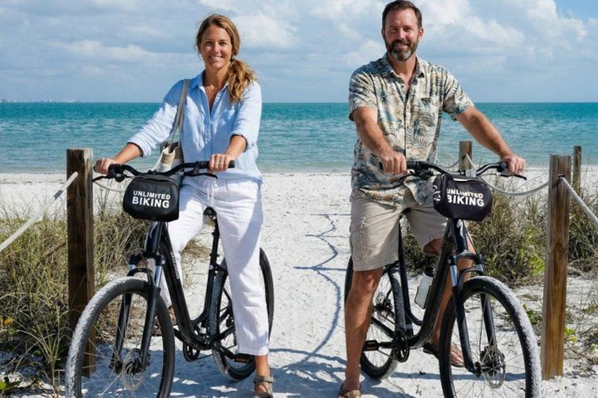 Two people on bicycles with 'UNLIMITED BIKING' bags, a sandy beach, and the ocean in the background. 2