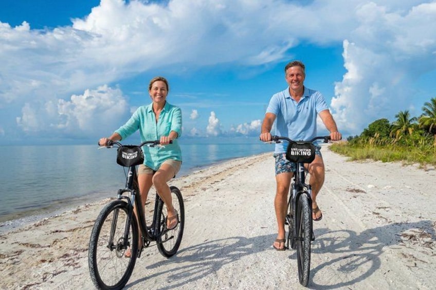 Two people riding bicycles on a sandy beach with the ocean in the background. 1