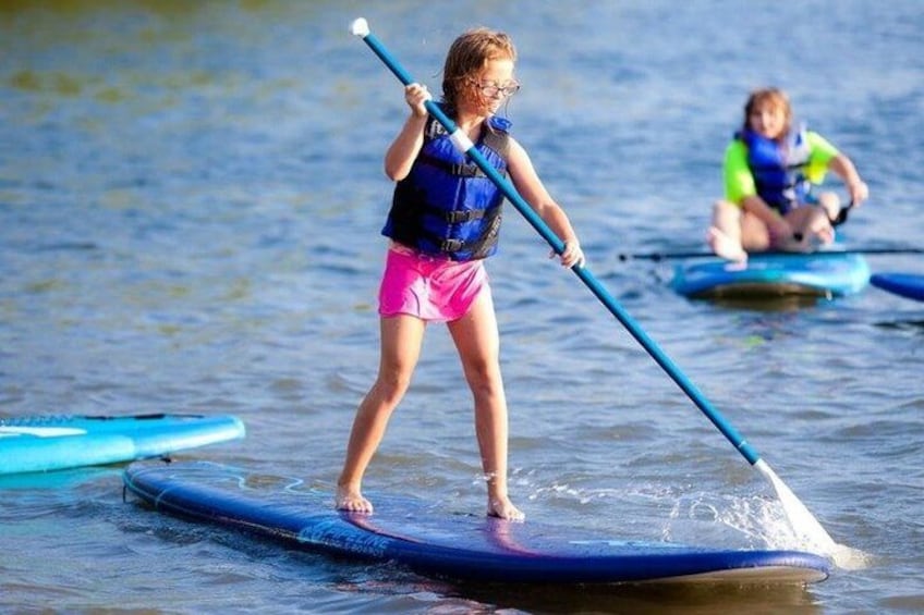 A child paddleboarding on a calm body of water, wearing a life jacket and pink shorts. 3