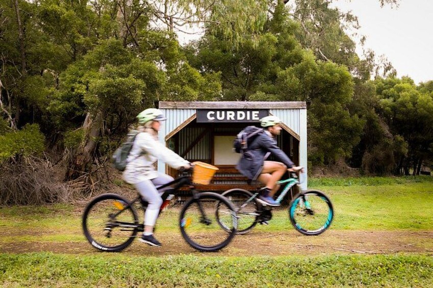 Two cyclists riding bicycles on a dirt path with a small wooden structure labeled 'CURDIE' in the background. 1