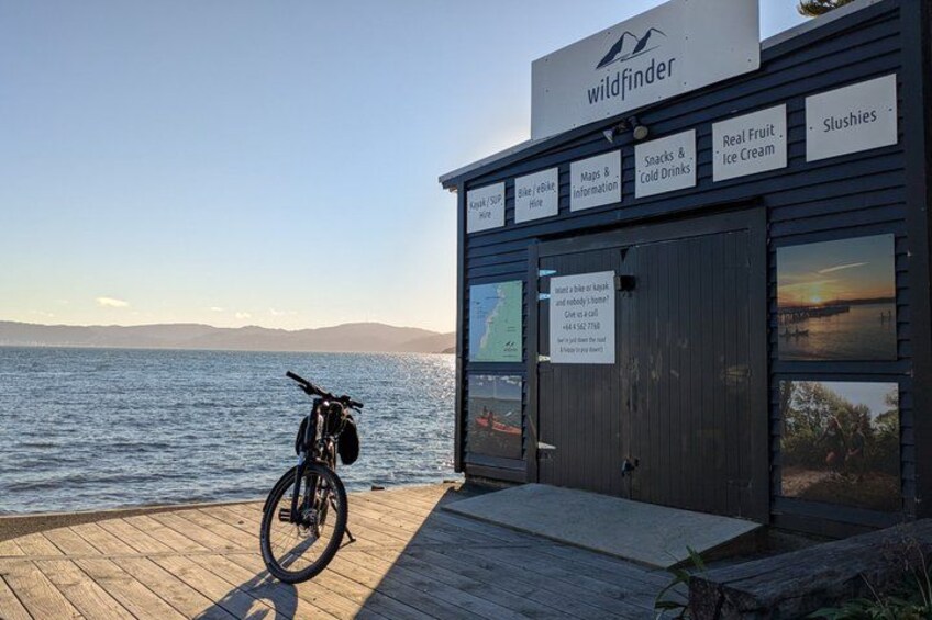 A wooden dock with a bicycle, a building labeled 'wildfinder' with signs for maps, snacks, and ice cream, and a view of the water and mountains in the background. 2
