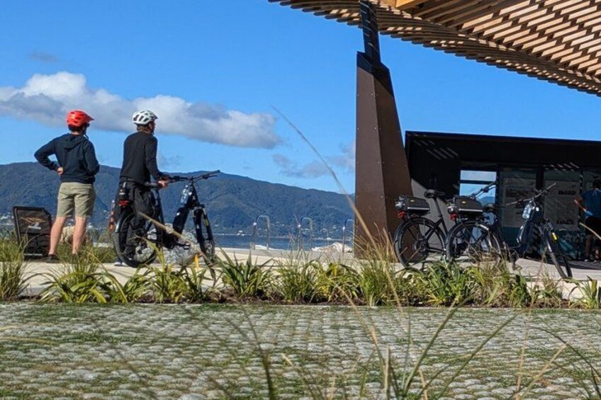 Two people standing near bicycles, a building with a sloped roof, and mountains in the background. 1