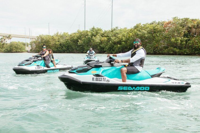 Two people riding jet skis on a body of water with a bridge and greenery in the background. 1