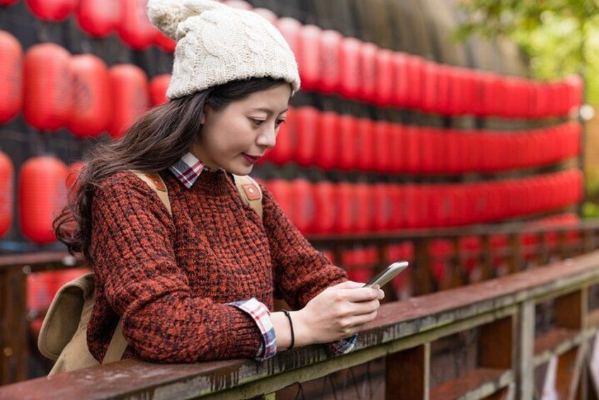 A person sitting on a wooden railing, wearing a knitted sweater and a white hat, holding a smartphone. 1