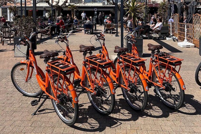 A row of orange bicycles parked on a paved area with people sitting at tables in the background. 1