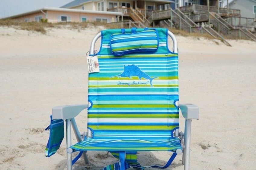 A beach chair with a striped cover and a matching bag, set on a sandy beach with a house and stairs in the background. 5