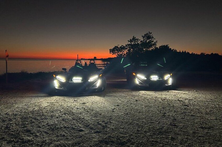 Two vehicles with headlights on, parked on a gravel surface near a body of water at sunset. 2