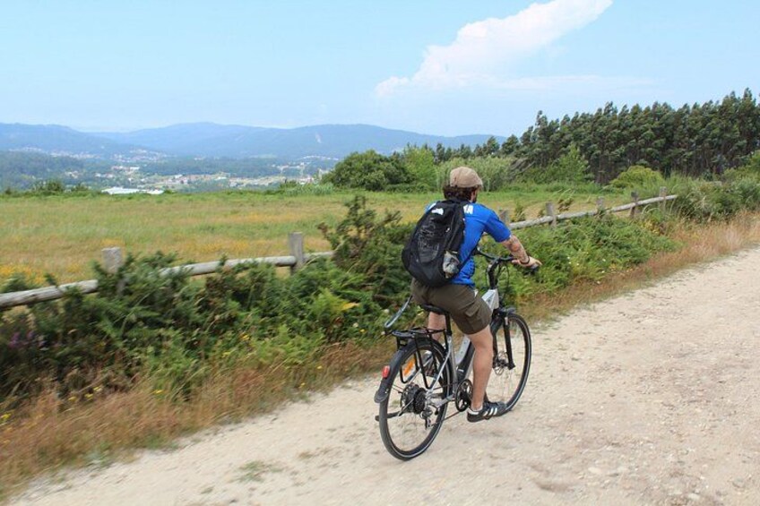 A person riding a bicycle on a dirt path with a backpack, surrounded by greenery and a distant view of a town. 3