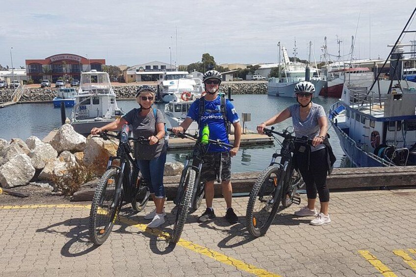 Three people on bicycles at a marina with boats and rocks in the background. 3
