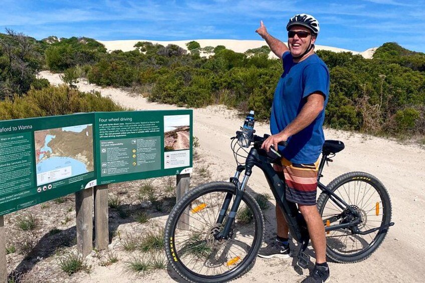 A man with a bicycle on a sandy trail, a signboard with information, and a clear sky. 5