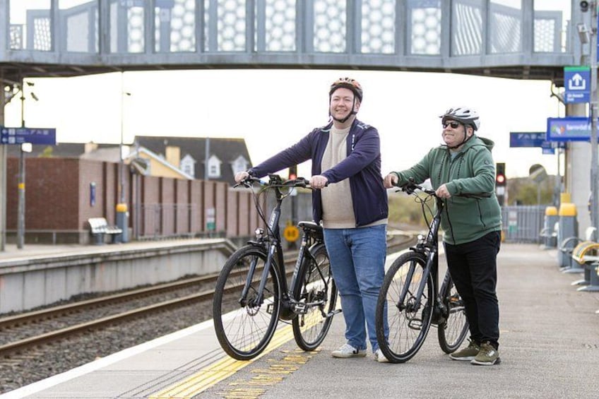 Two people standing on a train platform with bicycles, a bridge in the background, and a train track. 5
