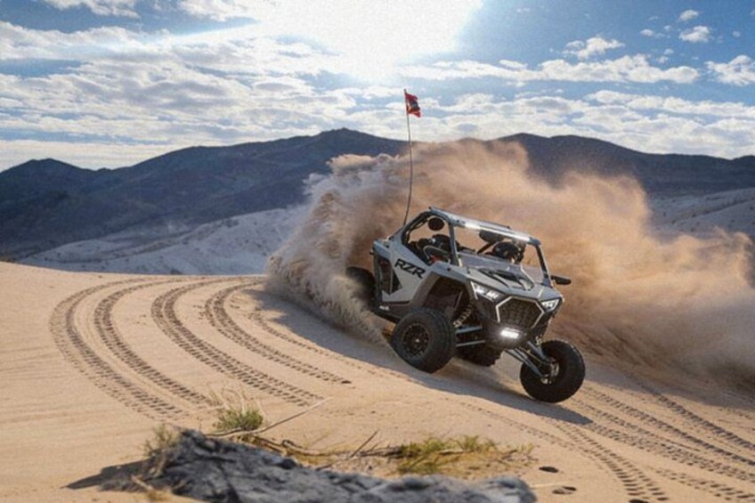 A white off-road vehicle with a flag, kicking up dust in a desert with mountains in the background. 1