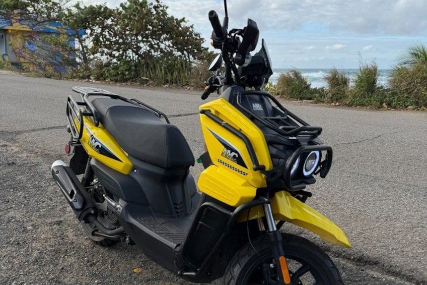 A yellow and black scooter parked on a paved road with a coastal background. 3