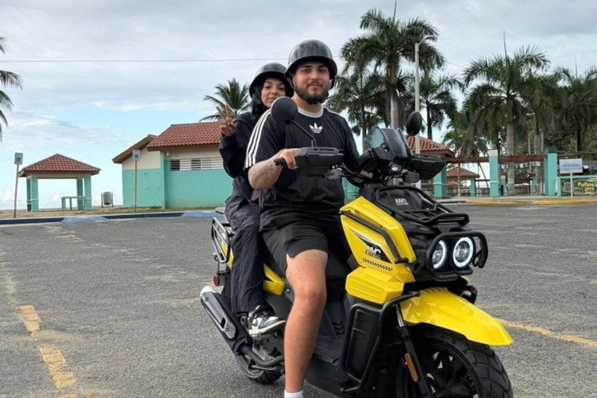 Two people on a yellow motorcycle in a parking lot with palm trees and buildings in the background. 5