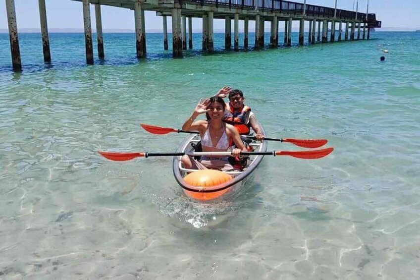 A kayak with two people paddling on clear turquoise water, a pier in the background, and a clear sky. 1