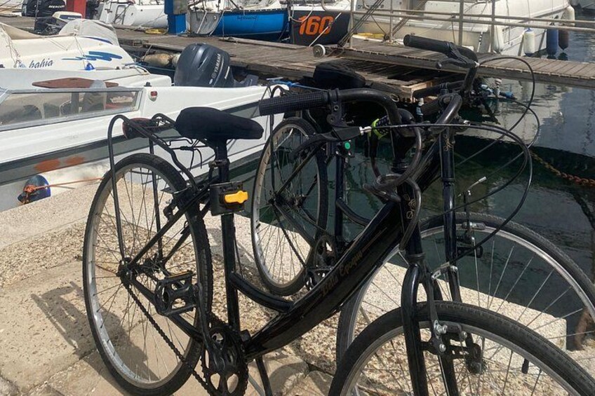 A black bicycle with a rear rack and a seat attached, parked on a concrete surface near a body of water with boats in the background. 4