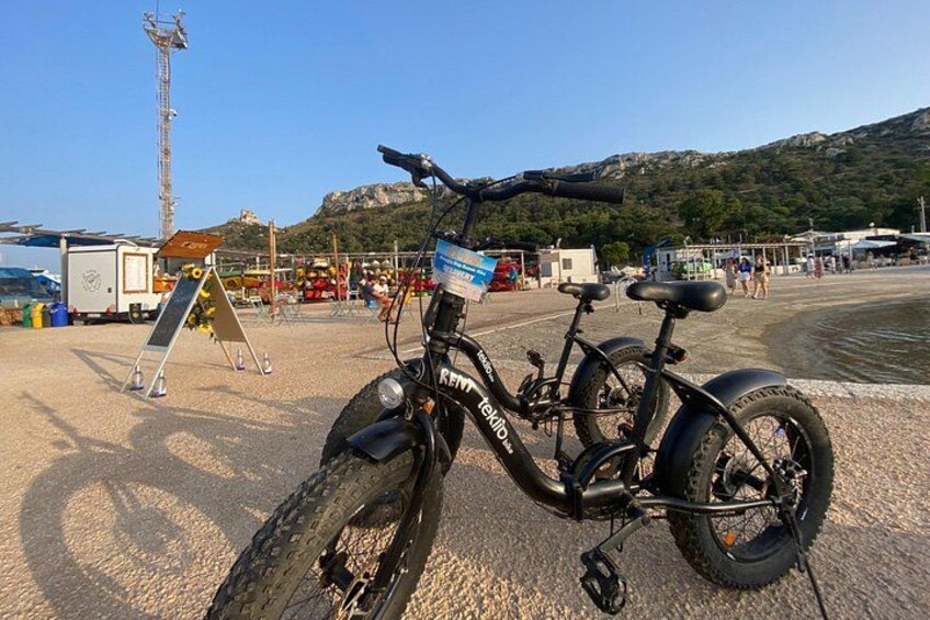 A black electric bicycle parked on a gravel surface near a body of water with mountains in the background. 2