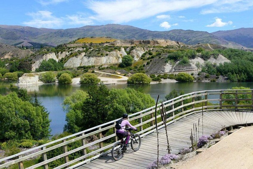 A person riding a bicycle on a wooden bridge over a river, surrounded by greenery and hills. 3