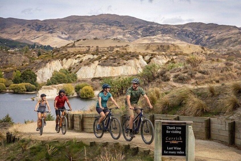 Four cyclists on a dirt path with a sign that reads 'Thank you for visiting Carrick Last Wine for many miles.' 1