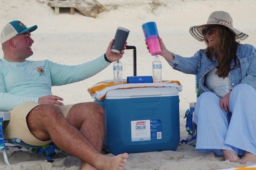 Two people sitting on beach chairs with a cooler and drinks. 5