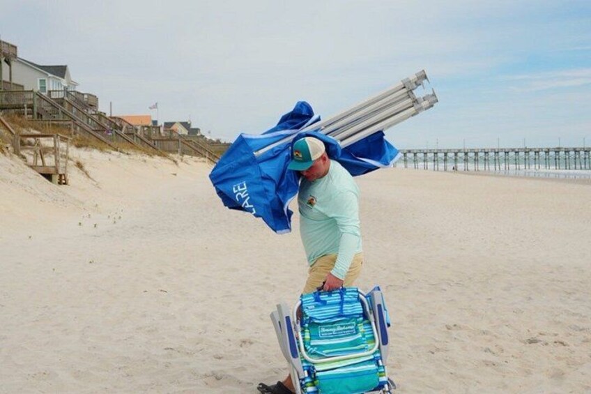 A person carrying a blue bag and a blue umbrella on a beach with a wooden walkway and a pier in the background. 2