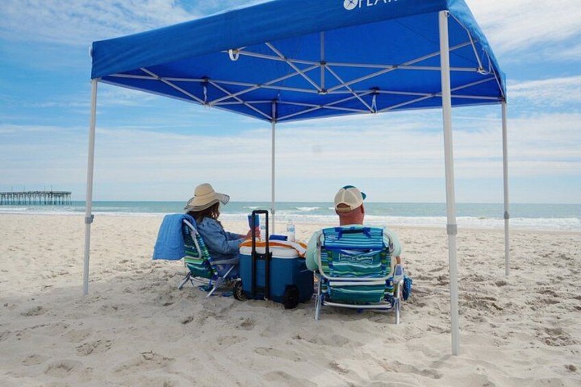 Two people sitting under a blue canopy on a sandy beach with a pier in the background. 3
