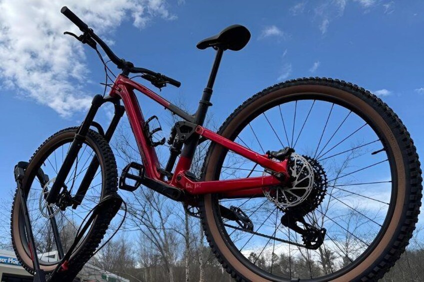 A red mountain bike with a front suspension fork, a rear derailleur, and a rear cassette, leaning against a large mountain bike tire. 2