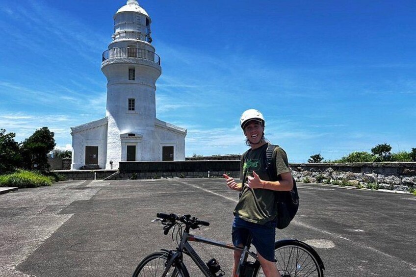 A person standing next to a bicycle in front of a white lighthouse. 1
