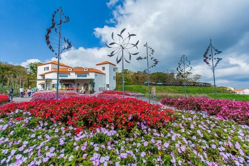Ein Haus mit rotem Dach, umgeben von bunten Blumenbeeten und dekorativen Windspielen. 1