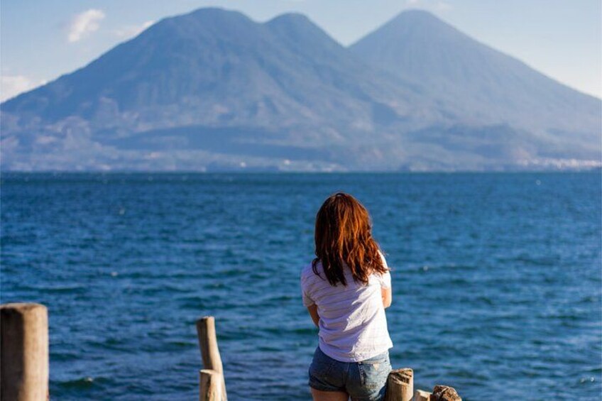 A person sitting on a wooden structure by the sea, facing a mountain range. 1