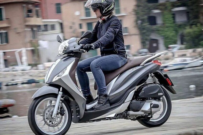 A person riding a scooter on a paved path with buildings and water in the background. 1