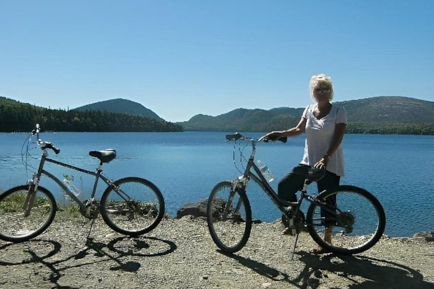 A person standing next to a bicycle by a lake with mountains in the background. 2