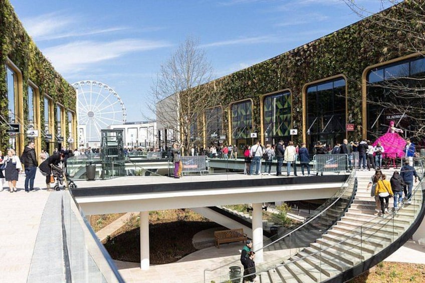 A pedestrian bridge with glass railings, a Ferris wheel in the background, and a modern building with large windows. 2