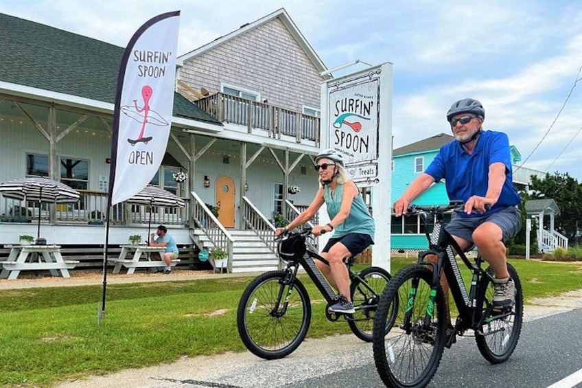 Two people on bicycles in front of a building with a sign that reads 'SURFIN' SPOON OPEN'. 2