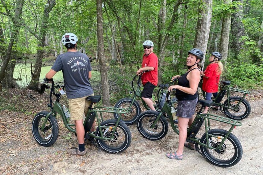 A group of people on electric bikes in a forested area with trees and a dirt path. 4