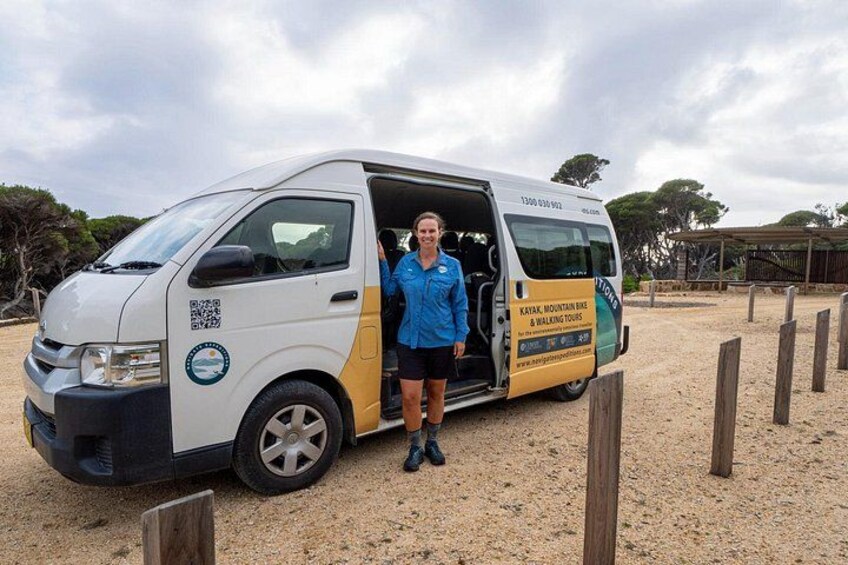 A white and yellow van with a QR code and text on the side, parked on a gravel surface with wooden posts in the background. 1
