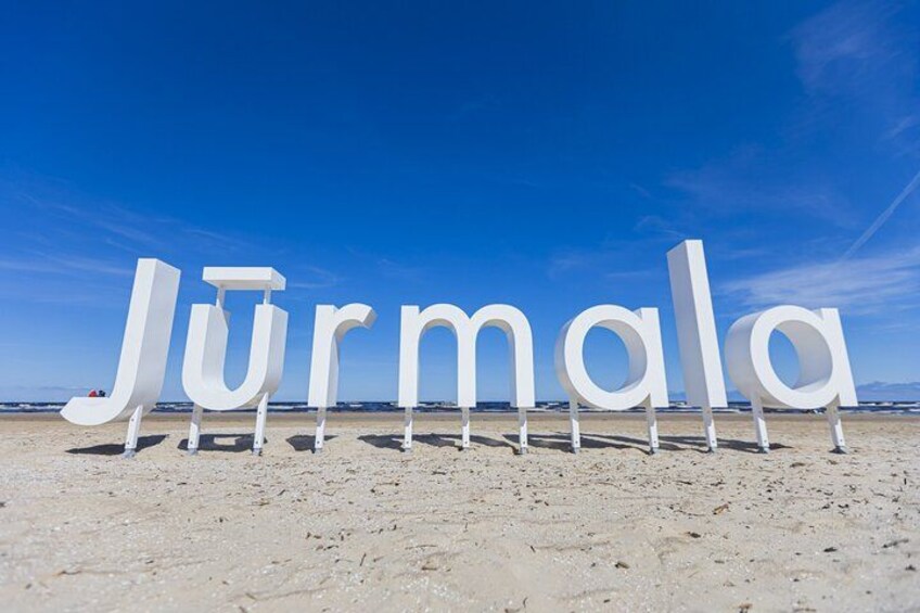 Large white letters spelling 'Jurmala' on a beach with a clear blue sky. 1